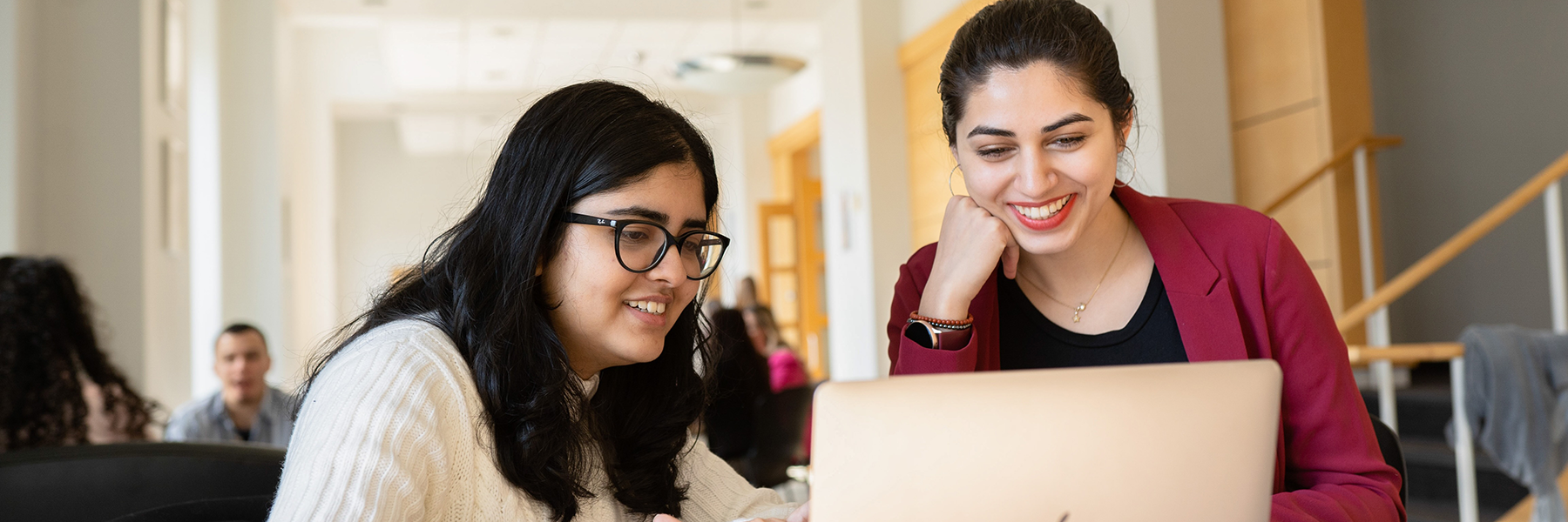 Two college-aged students, a woman with glasses and a woman with a burgundy blazer, sit side-by-side looking at a laptop computer. They are smiling and appear to be studying or collaborating on a school project in a bright, modern setting.